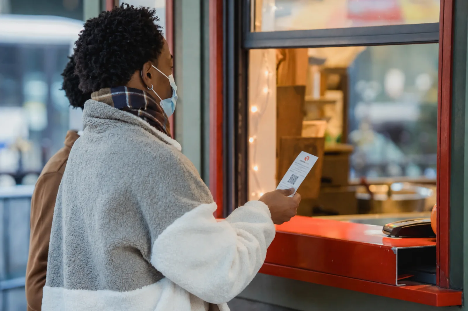 Gast hält am Verkaufsfenster einen gedruckten Gutschein-Bon mit QR-Code in der Hand